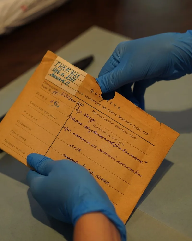Sobre de archivo de papel envejecido, con un desgarro en el borde superior, sostenido con guantes sobre una mesa de examen.