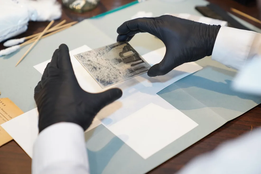 Manos con guantes sostienen una placa fotográfica sobre una mesa de trabajo con hisopos y hojas de papel de conservación.